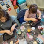 Zwei Schülerinnen sitzen an einem Tisch und haben jeweils eine schwarze Schale in der Hand. Auf dem Tisch neben ihnen liegen verschiedene Dekoartikel wie Bänder und Farben. In den Schalen, welche sie halten, befindet sich eine körnige Masse, ähnlich wie große Salzkristalle.