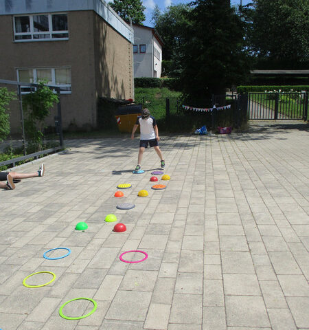 Auf dem Boden liegen verschiedene Dinge wie Reifen oder Balancepads in einer Reihe. Am Anfang steht ein Schüler, der in die Reifen und auf die Balancepads springt, um die Reihe zu schaffen.