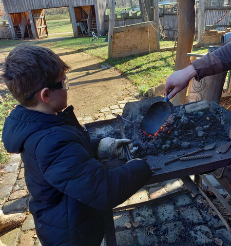 Das historische Dorf in Zwickau. Zu sehen ist ein kleiner Schüler, welche eine Schutzbrille, Lederhandschuhe und einen langen Anorak trägt. Er steht mit einem Erwachsenen am Schmiedeofen und hält mit einer langen Zange ein Werkstück fest, während der Erwachsene dieses mit einer Schaufel mit glühenden Kohlen bedeckt.